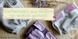 Ein Foto von handgemachten Stoffwindeln in neutralen Grau- und Beerentönen, verziert mit Knöpfen und Bändchen im traditionellen bayerischen Stil, angeordnet auf einem Holzuntergrund mit Stroh und blauen Blumen im Hintergrund. Über den Windeln steht in geschwungener Schrift "Stoffwindeln aus Bayern – so wicklst gscheid!" und in der unteren rechten Ecke befindet sich ein Herz-Logo. Die Bildquelle "Windl fürs Kindl" ist am unteren Rand des Bildes angegeben.